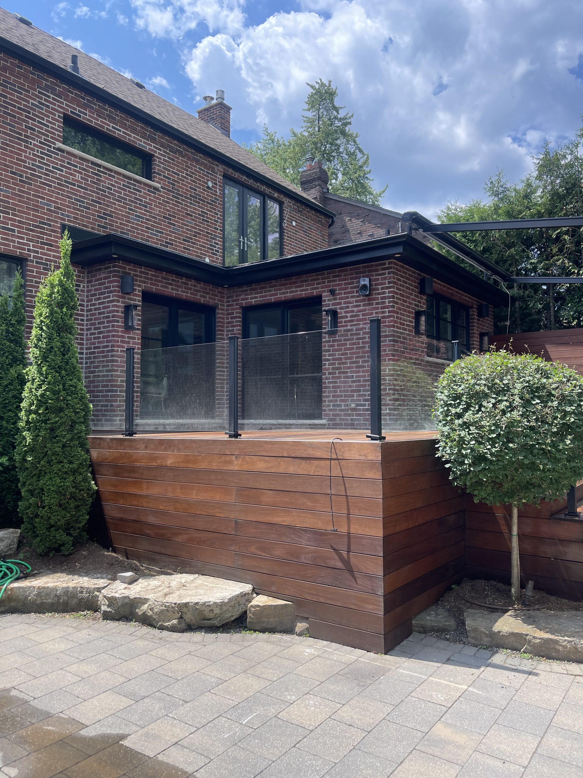 Exterior renovation showcasing a modern wooden deck with glass railing, brick house facade, and landscaped greenery, emphasizing Urban Reno Design's commitment to quality and aesthetic appeal.